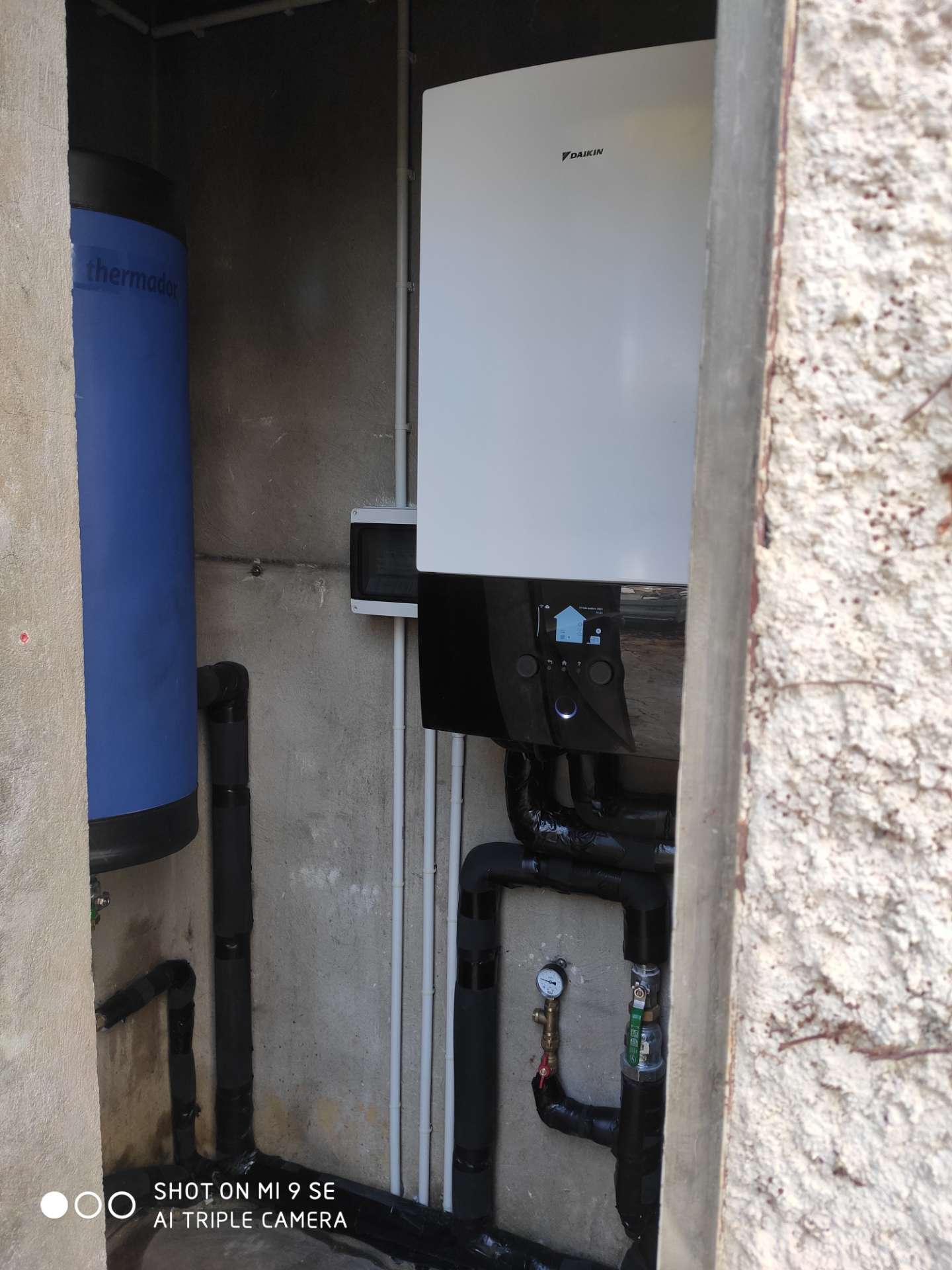 Un chauffe-eau mural blanc marqué DAIKIN au-dessus de tuyaux noirs isolés, à côté d'un ballon bleu marqué thermador, dans un local technique en béton.